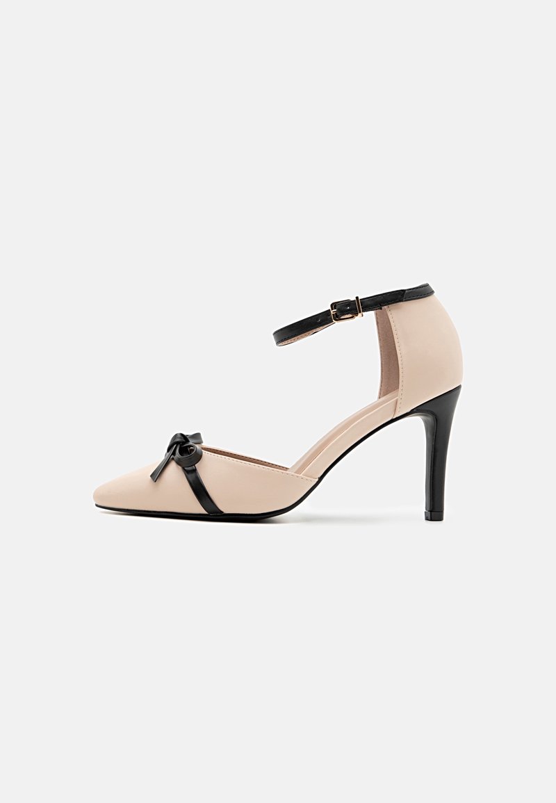 Chaussure beige à talon haut avec bout pointu, bride de cheville noire avec boucle, talon noir et détail de nœud noir à l'avant.