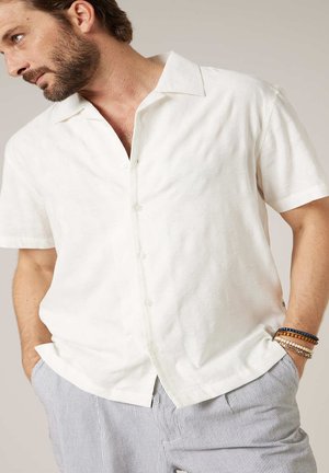 Man met baard die een wit overhemd met korte mouwen en knopen draagt en lichtblauwe gestreepte broek, met handen in de zakken en kralenarmbanden om zijn linker pols.