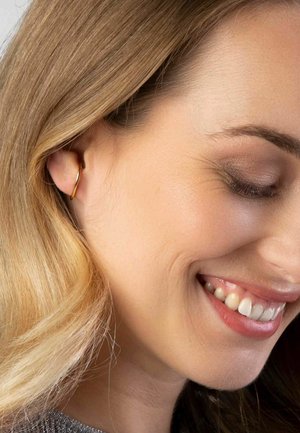Boucle d'oreille en or avec un design circulaire lisse, portée à l'oreille gauche. Les cheveux sont coiffés en vagues lâches, encadrant partiellement le visage.