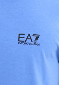 Lyseblå T-skjorte laget av mykt stoff, med en svart EA7-logo og teksten "Emporio Armani", satt mot en jevn tekstur.
