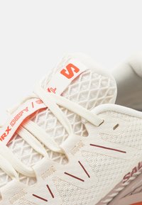 Lettvekts kremfarget joggesko med mesh-overdel, snøringslukking, røde logo-detaljer og teksturert design på sidene for bedre grep og ventilasjon.