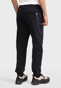 Pantalones jogger negros con cintura elástica, dos bolsillos traseros y una cadena decorativa plateada adjunta, combinados con zapatillas beige y negras.