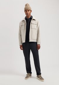 Light gray wool blend jacket with flap pockets, paired with a black polo shirt and dark pants. Brown sneakers with beige accents.