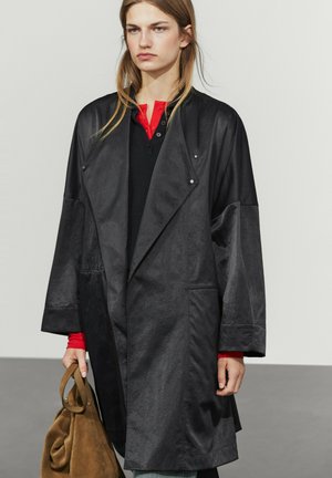Giovane con lunghi capelli indossa un cappotto nero ampio sopra una camicia rossa e tiene una borsa in camoscio marrone su uno sfondo grigio uniforme.