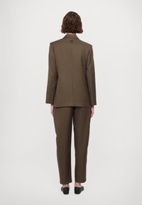 Bruine op maat gemaakte blazer met schoudervulling en een enkele rugventilatie, gecombineerd met bijpassende broeken. Gladde stof, minimalistisch ontwerp, slanke silhouet.