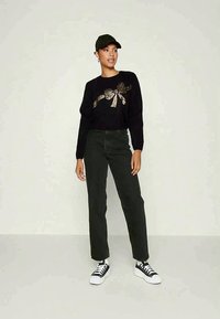 Czarny sweter z metalicznym wzorem w kształcie kokardy, ciemnozielone spodnie i czapka w kolorze czarnym. Model nosi białe sneakersy z czarnymi akcentami, stojąc na neutralnym tle.