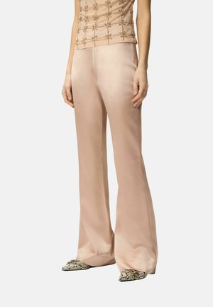 Žena oblečená v béžových širokých saténových nohaviciach, bezrukávovom vrchom zdobenom korálkami a v špicatých hadích topánkach, stojaca pred bielym pozadím.