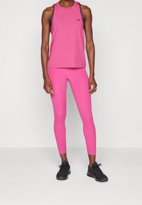 Różowy sportowy top na ramiączkach z logo na piersi i ozdobnymi paskami; w komplecie z dopasowanymi różowymi legginsami i czarnymi butami sportowymi. Gładka faktura.