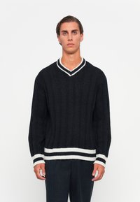 Jeune homme se tenant face à l'objectif, portant un pull en tricot noir avec des rayures blanches au col, aux poignets et à l'ourlet sur un fond uni.