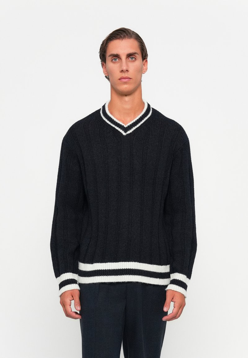 Jeune homme se tenant face à l'objectif, portant un pull en tricot noir avec des rayures blanches au col, aux poignets et à l'ourlet sur un fond uni.