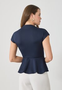 Top peplum azul marino con un diseño ajustado y suave, mangas cortas y un dobladillo con puntas. El material parece elástico y suave. Pendientes de aro dorados visibles.