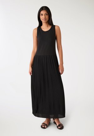Abito lungo nero con corpetto aderente e gonna plissettata. Tessuto liscio sulla parte superiore, pieghe strutturate nella gonna. Indossato con sandali neri con cinturini.