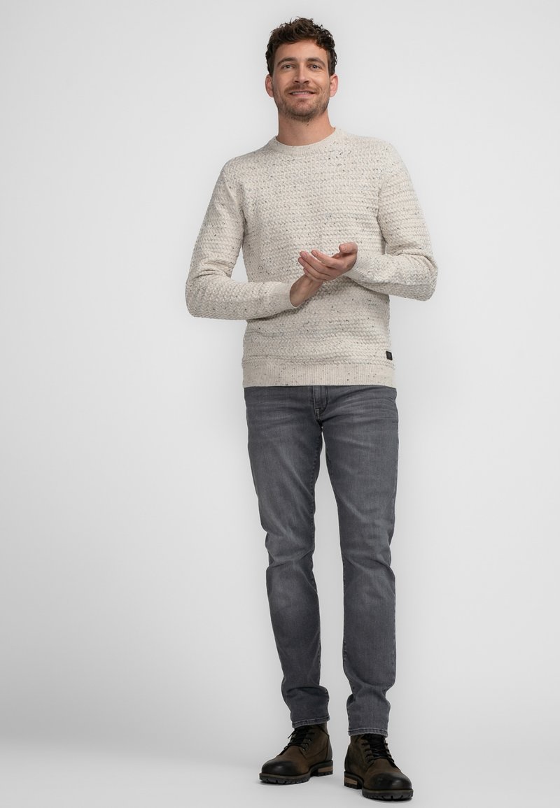Light gray knitted sweater with textured pattern, paired with dark gray jeans. The model wears brown lace-up boots and stands with hands clasped.