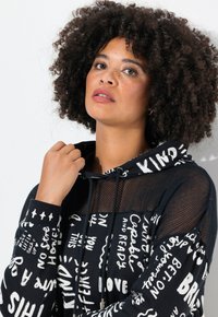 Sudadera negra con texto blanco y patrones gráficos, que presenta un detalle de malla en la capucha y las mangas. Tejido suave, corte relajado y bolsillo frontal.
