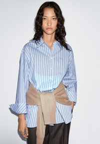 Blue striped shirt with a relaxed fit and oversized sleeves, paired with a beige sweater tied at the waist over dark pants.