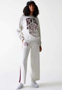 Completo felpa e pantaloni in cotone grigio chiaro con grafiche borgogna. I pantaloni hanno strisce laterali e tasche. Abbinati a sneakers bianche e un cappellino nero.