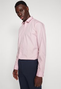 Chemise rose clair à manches longues avec un col pointu, fermeture à boutons sur le devant et détails aux poignets. Associée à un pantalon sombre.