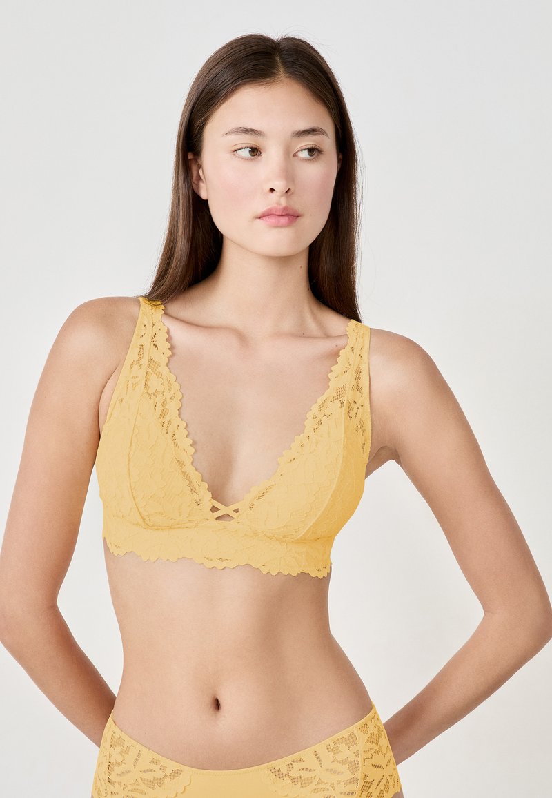 Bralette in pizzo di un giallo tenue, con scollatura profonda e bordi ondulati. Le spalline incrociate davanti aggiungono un tocco di dettaglio. Tessuto liscio.