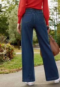 Jeans bleu à jambes larges avec une taille haute, deux poches arrière, ourlet légèrement usé, assortis à des baskets blanches et un sac à bandoulière marron.