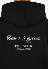 Čierna mikina s kapucňou so bielym vyšívaným textom: "Odváž sa byť iný," "PEGADOR" a "Šepoty chladu" na chrbte. Bavlnený materiál, bežný strih.