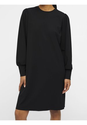 Woman wearing a simple black long-sleeve dress with puffed shoulders and cuffed sleeves, standing against a plain white background.