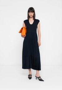 Marinblå midikjole med plisseringer, kroppsnær strikket overdel, cap sleeves og v-utringing. Modellen holder en oransje håndveske og har på seg svarte høye hæler.