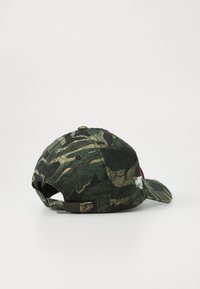 Groene en zwarte camouflagepet met verstelbare strap en een klein versleten label aan de zijkant, gezien van achteren op een witte achtergrond.