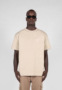 Beige cotton t-shirt featuring short sleeves and a crew neckline, with minimal white text detail on the front. Smooth texture.