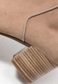 Beżowy zamszowy botek za kostkę z wzorzystym, tanowym obcasem w kształcie bloku oraz szwem wzdłuż górnej krawędzi. Gładka gumowa podeszwa.