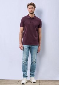 Polo à manches courtes, couleur bourgogne, avec un col, associé à un jean en denim bleu clair et des baskets blanches. Texture lisse et design ajusté.