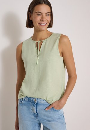Femme portant un haut vert clair sans manches avec un petit nœud devant, souriante, une main dans la poche d'un jean bleu clair.