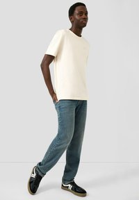 Jasnobeżowa koszulka z bawełny o okrągłym dekolcie, zestawiona z niebieskimi dżinsami. Czarne trampki z białymi akcentami dopełniają stylizację.