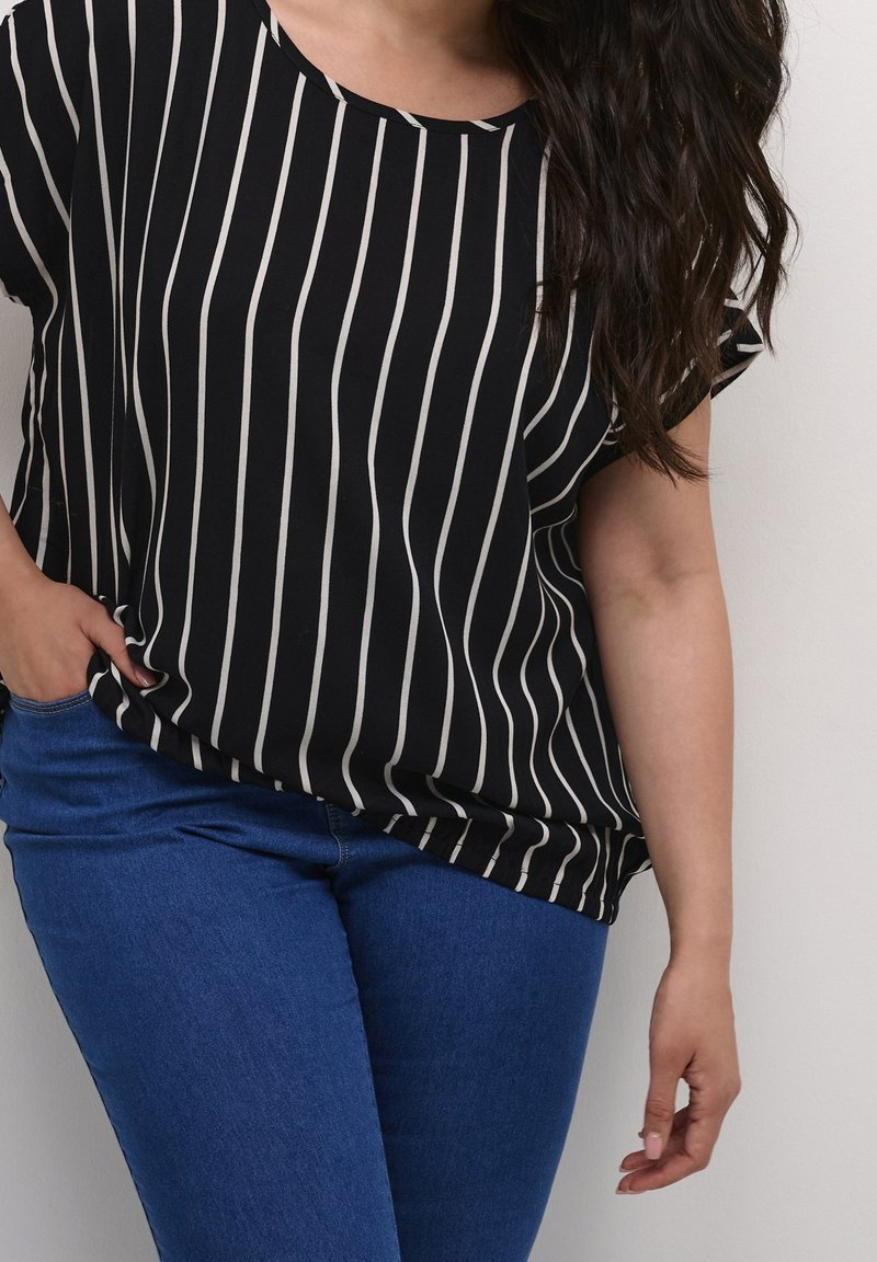 Femme portant une chemise rayée verticale noire et blanche et un jean bleu, une main dans la poche et de longs cheveux foncés sur l'épaule.