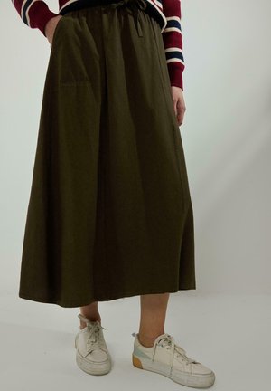 Oseba, oblečena v olive zelena midi krilo z vezalko v pasu in bele superge, stoji ob ozadju svetle, enobarvne stene.