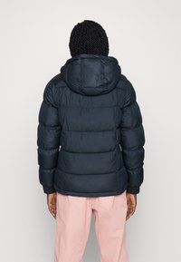 Námořnická puffer bunda s kapucí, se štepovaným designem a elastickými manžetami. Nositá přes světle růžové kalhoty, zobrazená ze zadní strany.