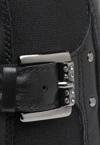 Primo piano di un cinturino in pelle nera con una fibbia in metallo argento decorata con piccole pietre brillanti trasparenti su un tessuto nero testurizzato.