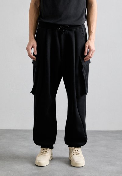 Pantalones de chándal negros, holgados, con bolsillos laterales y una cinturilla con cordón, combinados con zapatillas beige. Tejido suave y diseño relajado.