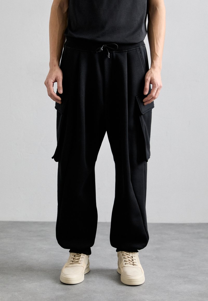 Black, loose-fitting sweatpants with side pockets and a drawstring waistband, paired with beige sneakers. Smooth fabric and relaxed design.