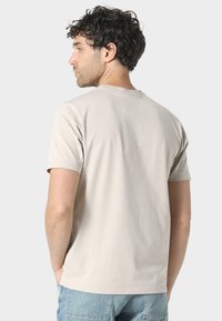 T-shirt in cotone beige chiaro con maniche corte e scollo rotondo, caratterizzato da una texture liscia e un design pulito e semplice sul retro.