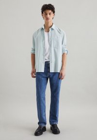 Lichtblauw gestreept overhemd met knopen over een wit T-shirt, gecombineerd met blauwe spijkerbroek en zwarte schoenen, staande tegen een neutrale achtergrond.