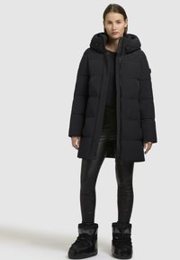 Lang sort pufferjakke med hætte, der har en quiltet design, lynlåslukning foran og trykknapper. Båret over sorte teksturerede leggings og kraftige sorte støvler.