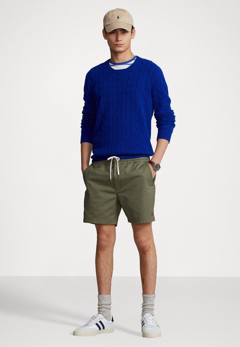 Blauer, strukturiert gestrickter Pullover, olivgrüne Shorts, graue Socken, weiße Sneakers mit marineblauen Streifen und eine beigefarbene Mütze mit einem Logo.