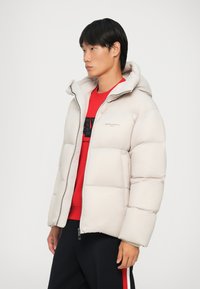 Lätta beige pufferjacka med dragkedja framtill, huva och quiltat mönster. Matchas med en röd sweatshirt och mörka byxor med logotyp.