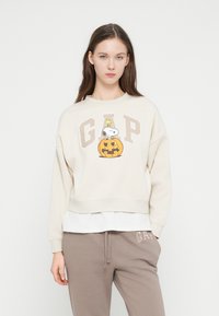 Φούτερ εκρού με εκτυπωμένο το Snoopy και γραφικό κολοκύθας, με το λογότυπο "GAP" σε μπεζ. Εμφάνιση με στρωμάτωση που συνδυάζει λευκό πουκάμισο από κάτω.