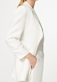 Biała marynarka z opadającym przodem, długimi rękawami i bocznymi kieszeniami. Gładki, lekki materiał o subtelnym połysku. Dopasowany krój.