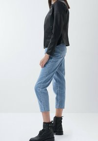 Donna con giacca di pelle nera, jeans corti blu chiaro e stivali alla caviglia neri con lacci e fibbie, in posizione laterale su sfondo bianco.