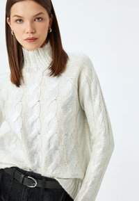 Kabelstrickpullover in zartem Creme mit hohem Kragen, der ein strukturiertes Muster und einen gerippten Saum aufweist. Stilisiert mit einem dunklen Gürtel und schwarzen Hosen.