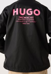 Czarna, lekka kurtka z dużym, różowym logo "HUGO" na plecach, a także z nadrukowanymi znakami towarowymi oraz datą założenia.