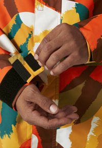Bunte Jacke mit orangefarbenen, gelben, grünen und braunen abstrakten Mustern. Nahaufnahme eines Handgelenks mit einem Klettverschluss und hochgekrempeltem Ärmel.