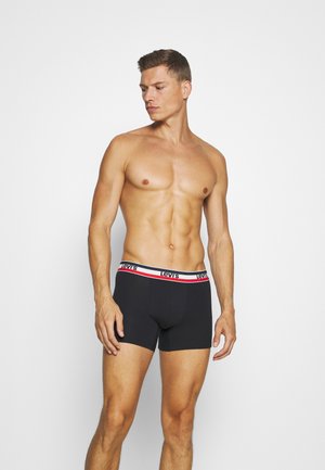 Schwarze Boxer-Briefs mit einem rot-weißen Bündchen, das das Levi's-Logo trägt. Das Modell steht oben ohne und zeigt eine muskulöse Figur.